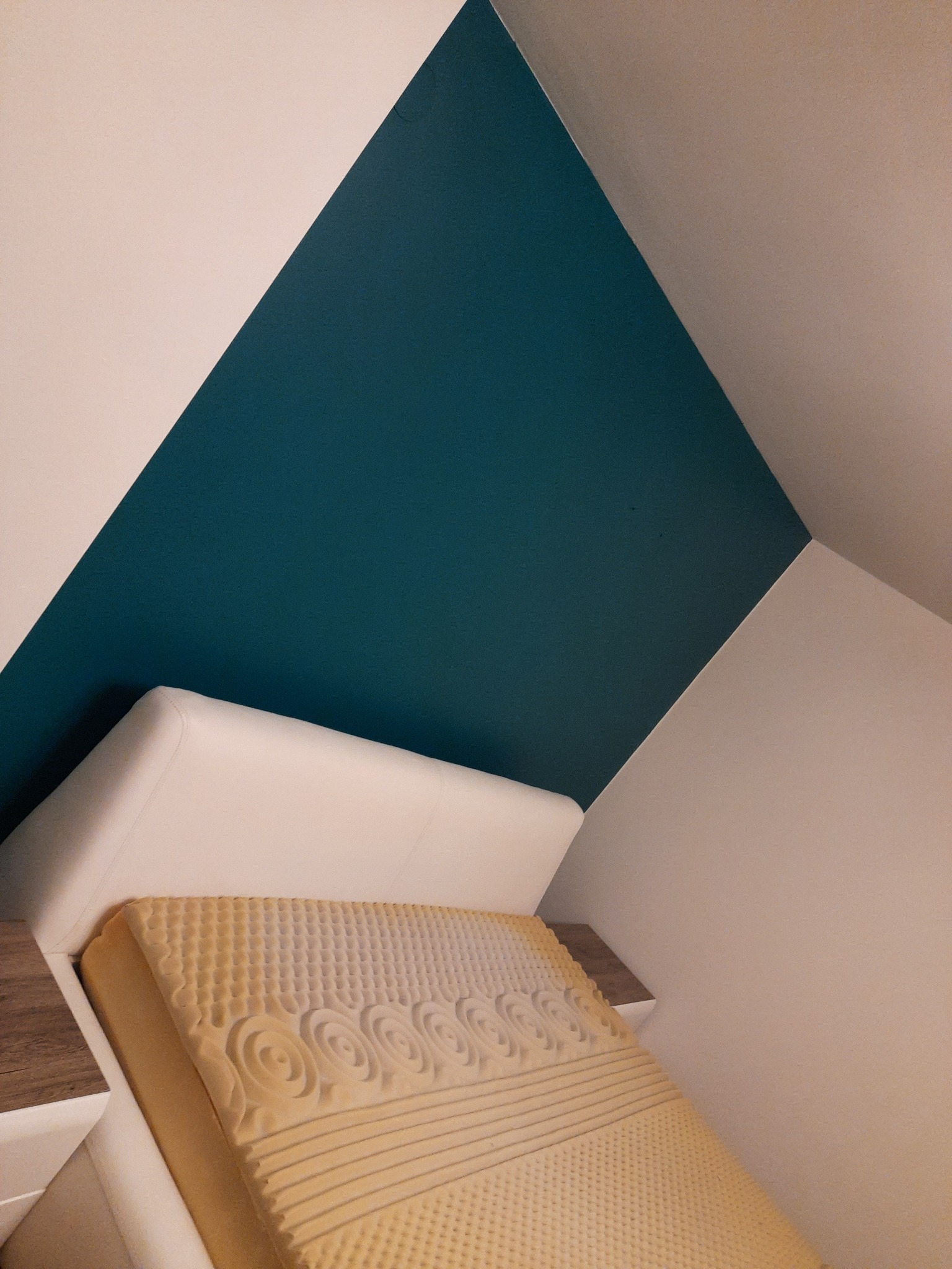 Pomieszczenie na poddaszu z ukośnym sufitem, gdzie ściana pomalowana jest w dwie barwy: białą i turkusową, pod ścianą stoi rozłożony tapczan z materacem z pianki.