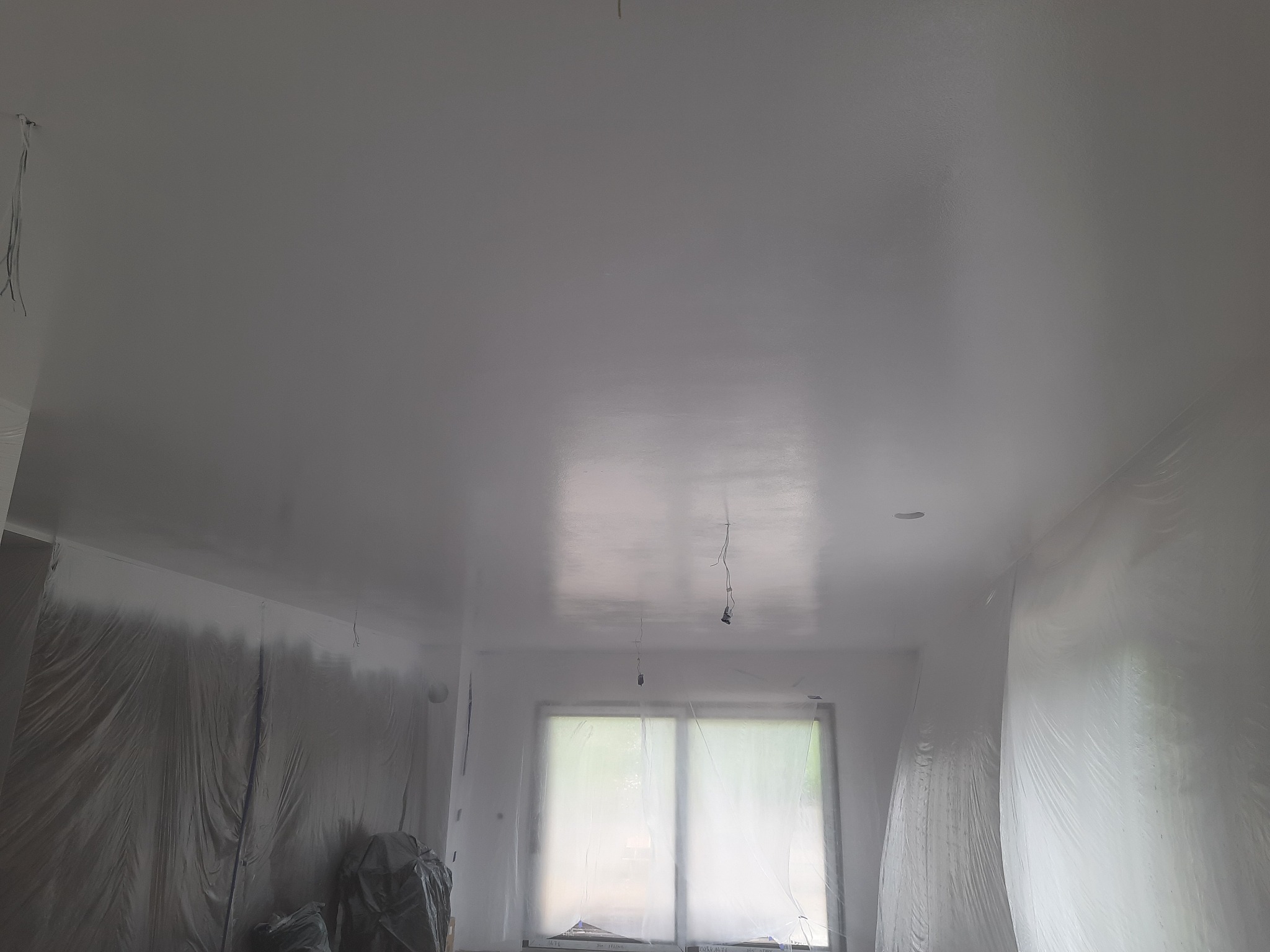 Białe, świeżo pomalowane wnętrze z zabezpieczonymi folią ścianami i oknem, widoczne wiszące przewody elektryczne.