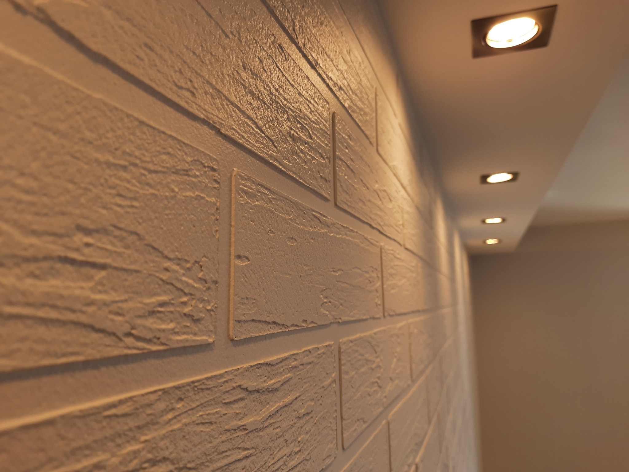 Ściana z dekoracyjną fakturą imitującą cegłę, oświetlona punktowymi lampami sufitowymi, z widoczną perspektywą i ciepłą tonacją.