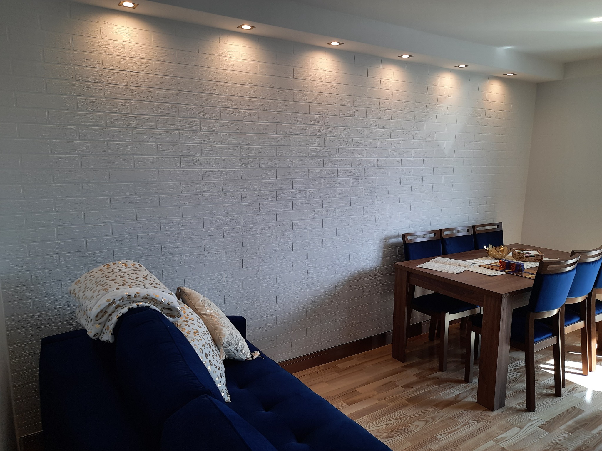 Biała ściana z fakturą cegły podświetlona punktowymi lampami sufitowymi w pokoju z drewnianą podłogą, niebieską sofą i stołem z krzesłami.