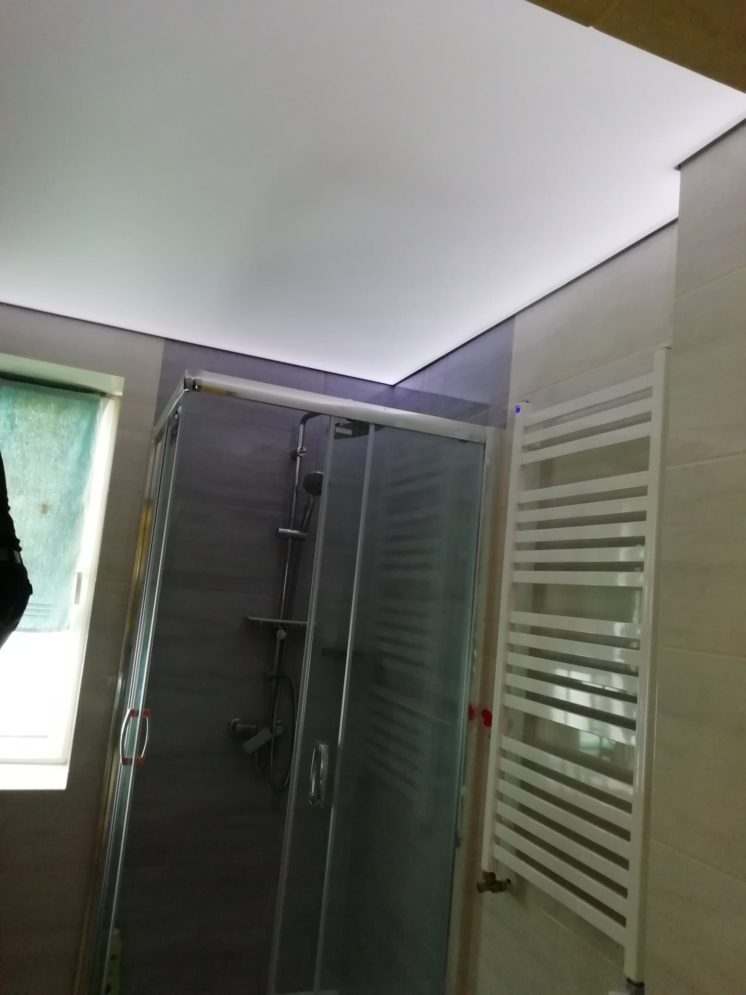 Nowoczesna łazienka z przeszkloną kabiną prysznicową i białym grzejnikiem drabinkowym, oświetlenie LED pod sufitem napinanym.