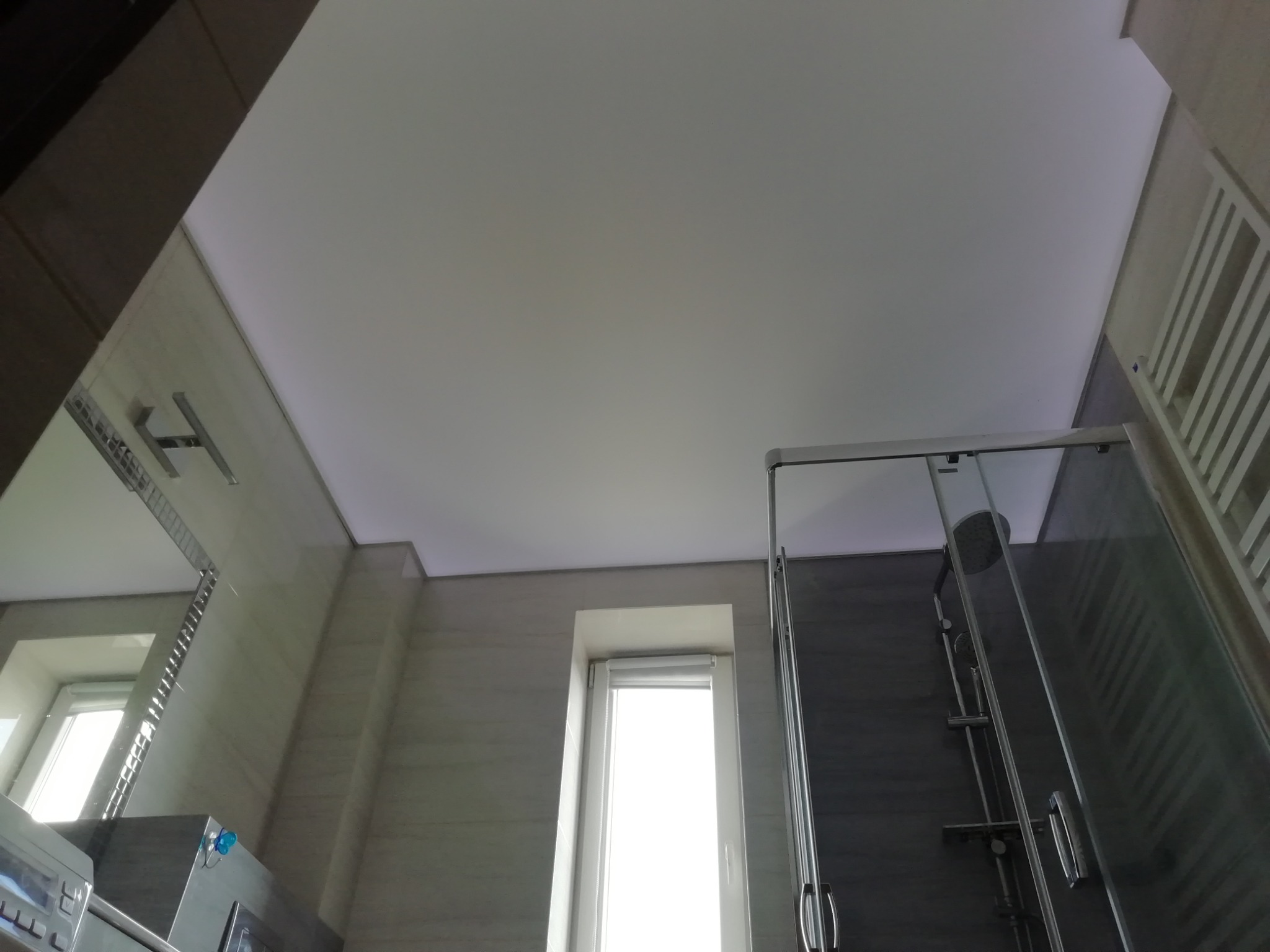 Biały, matowy sufit napinany w łazience z kabiną prysznicową i oknem, widok z dołu.