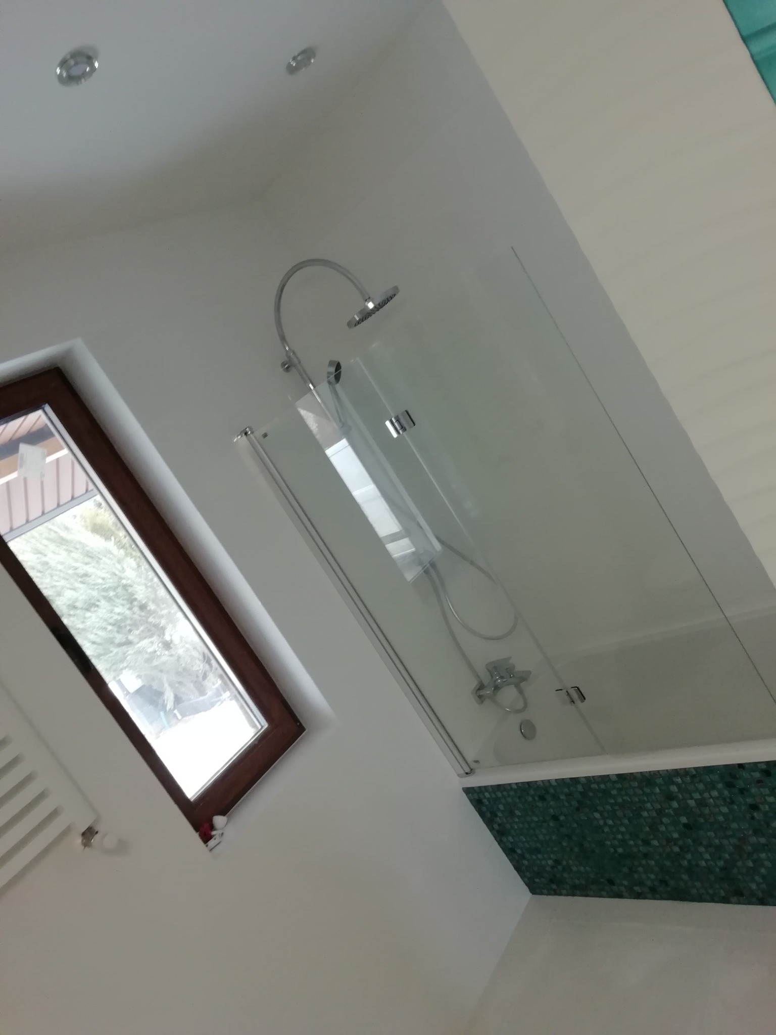 Nowoczesna łazienka z kabiną prysznicową z przezroczystego szkła, wykończona mozaiką w odcieniach zieleni, z oknem dachowym w drewnianej ramie i białymi ścianami.