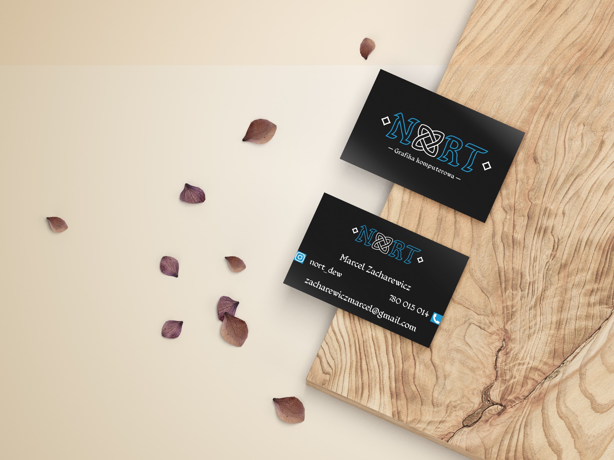 Dwie czarne wizytówki z logo 'N&RT - Grafika komputerowa' na drewnianej desce, otoczone luźno rozrzuconymi, suszonymi płatkami kwiatów.