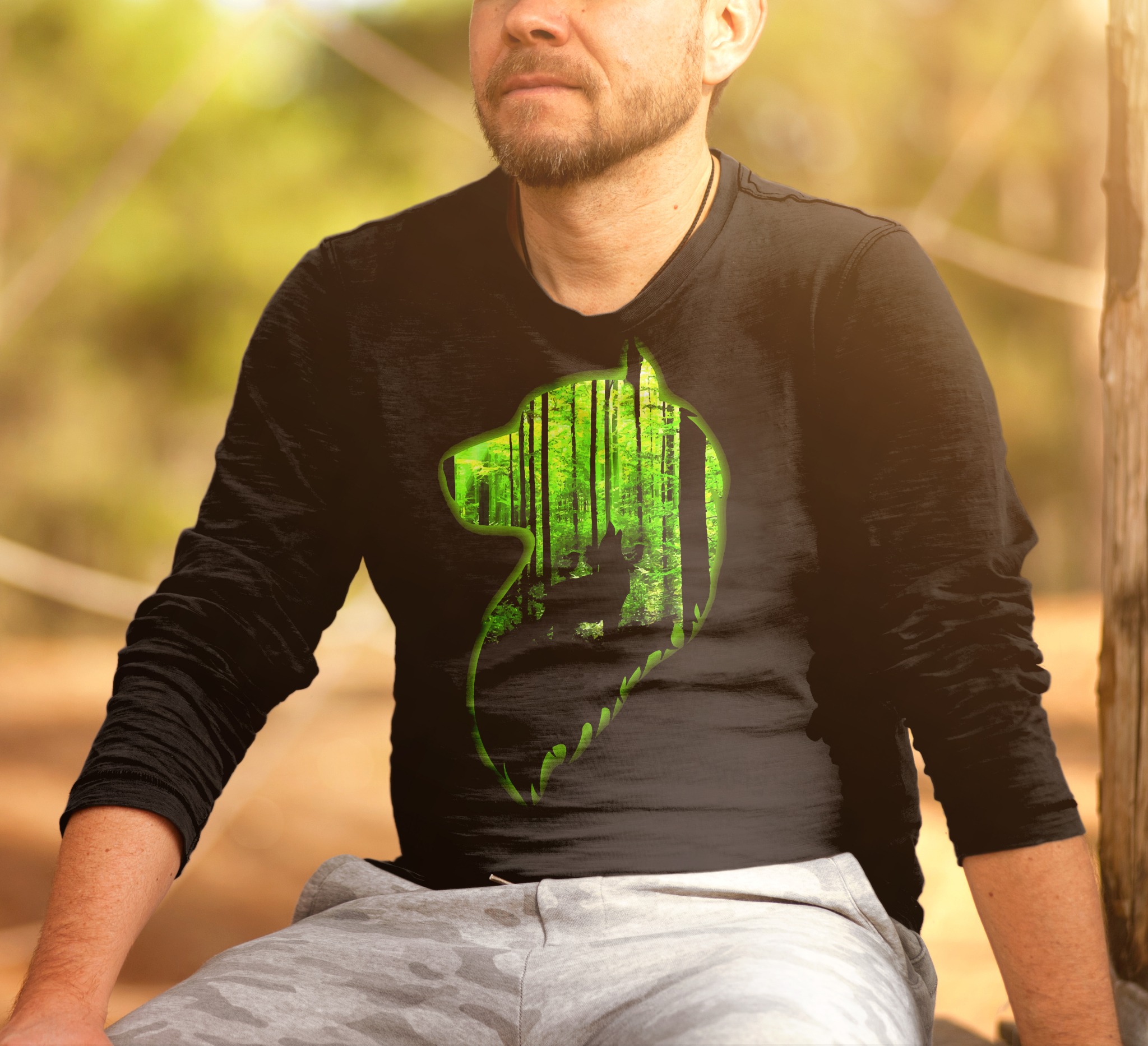 Mężczyzna w czarnej koszulce z długim rękawem, na której widnieje grafika przedstawiająca sylwetkę wilka wypełnioną motywem leśnym, siedzący na drewnianym pniu.