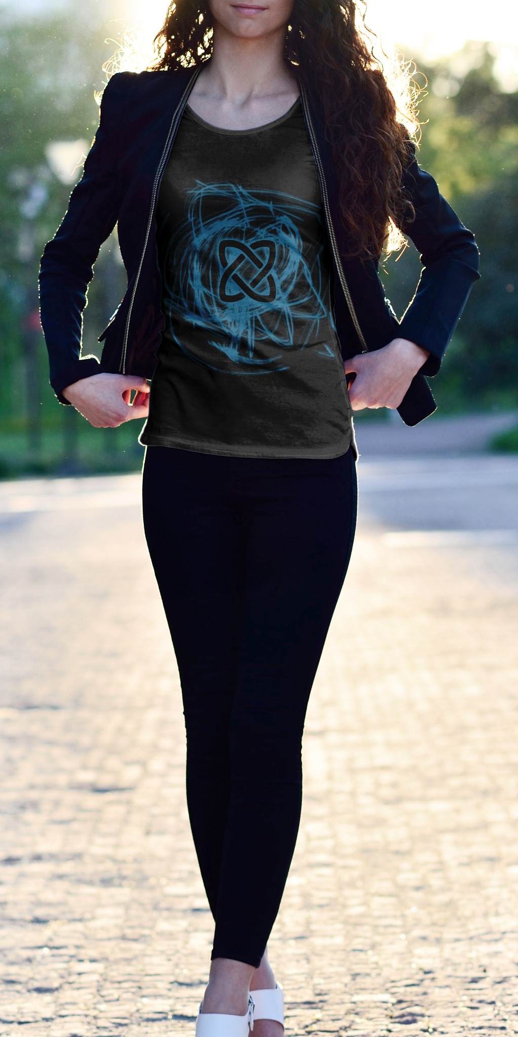 Kobieta w czarnym stroju prezentuje ciemną koszulkę z niebieskim, abstrakcyjnym wzorem celtyckiego węzła na tle słonecznej alejki.