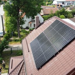 FORP Fotowoltaika i systemy grzewcze - Dwa panele fotowoltaiczne zainstalowane na brązowym dachu z dachówki ceramicznej, widok z góry na dom jednorodzinny i ulicę z drzewami w tle.