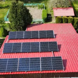 Widok z góry na panele słoneczne zamontowane na czerwonym, falistym dachu budynku, z ogrodem i małą szopą w tle.