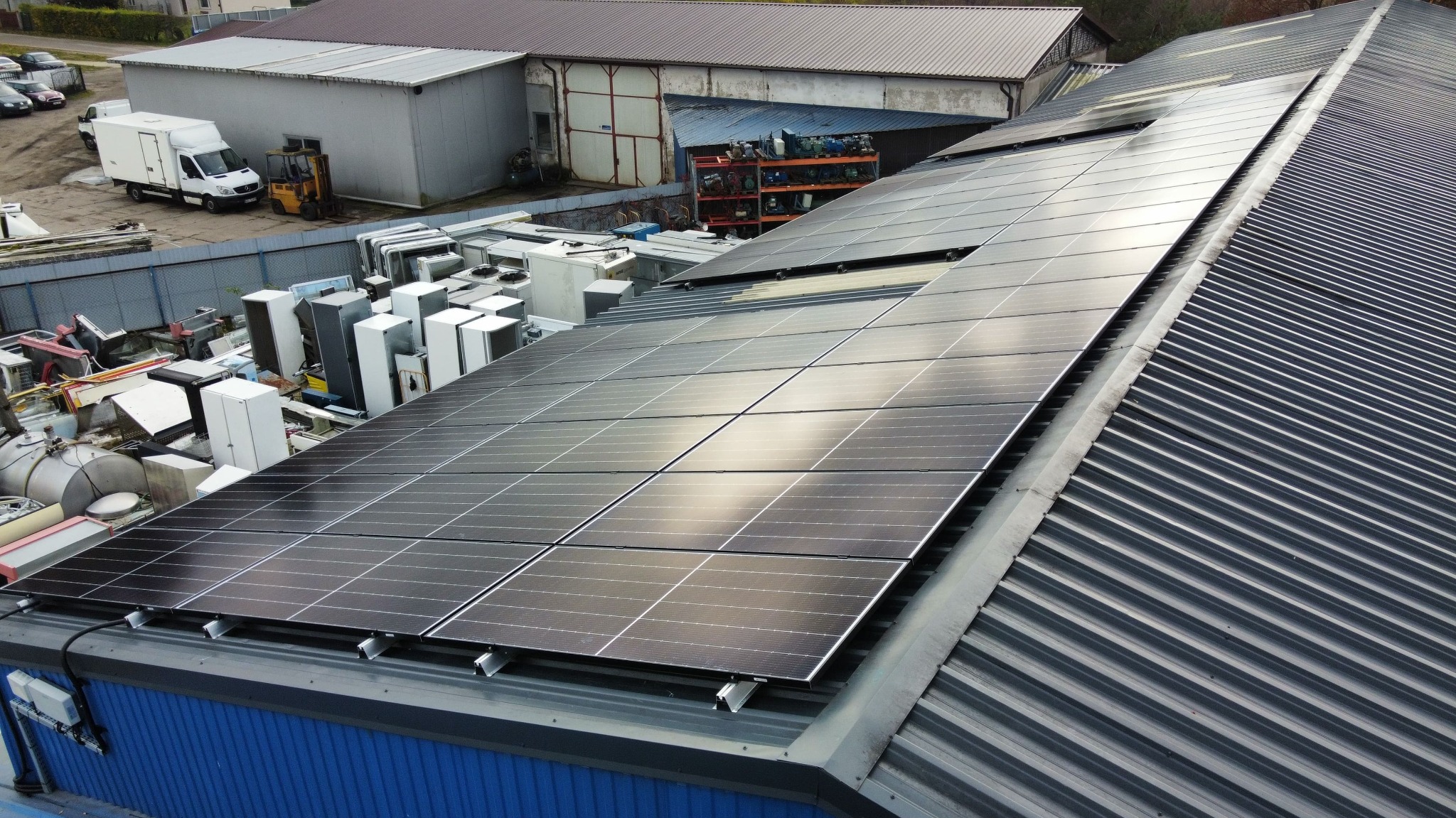 Panele słoneczne na dachu budynku przemysłowego z blachy trapezowej, widoczne odbicia światła i otoczenie magazynowe z pojazdami w tle.