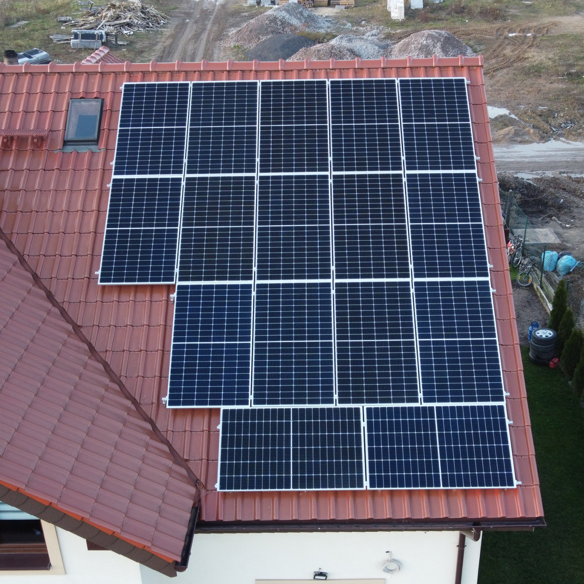 Widok z góry na panele słoneczne zamontowane na dachu z czerwonej dachówki w Olsztynie, z widocznym oknem dachowym i elementami otoczenia w tle.