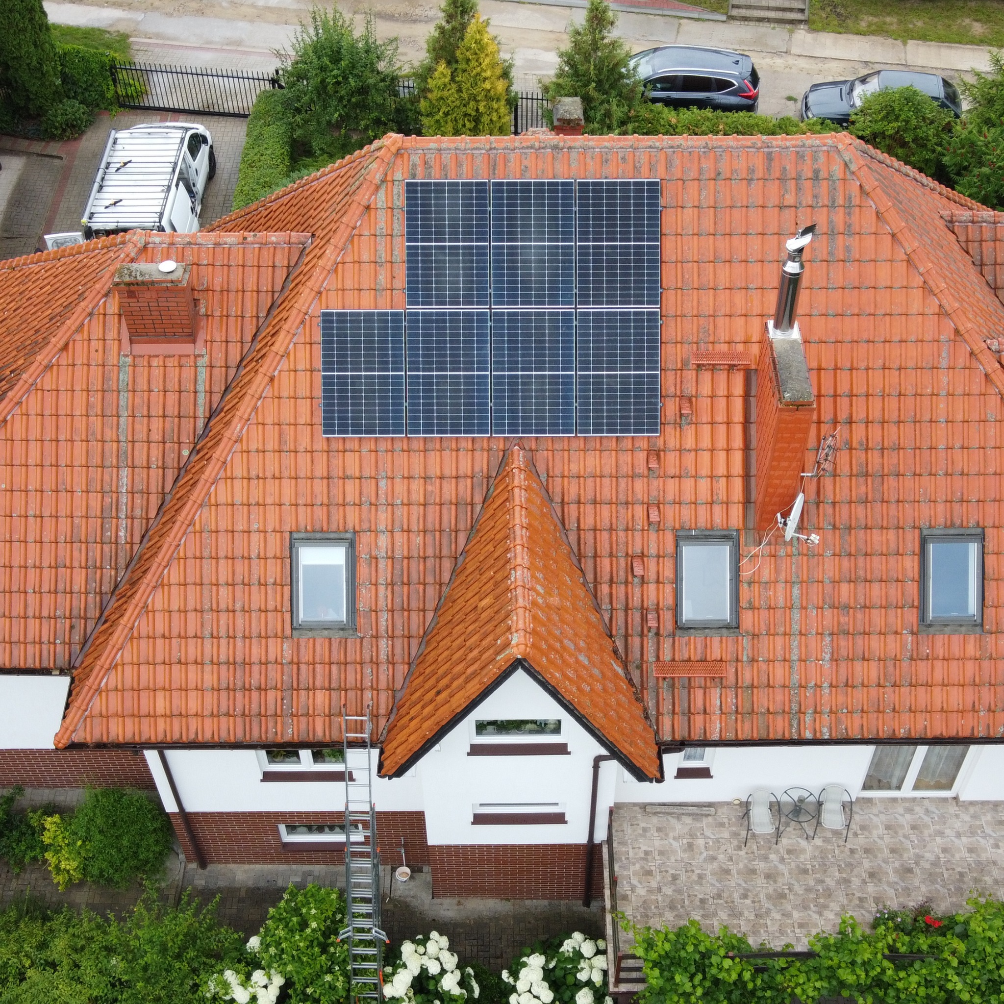 Widok z góry na dom z panelami słonecznymi na pomarańczowym dachu z dachówki. Przystawiona drabina, biały samochód dostawczy i zadbana zieleń wokół.