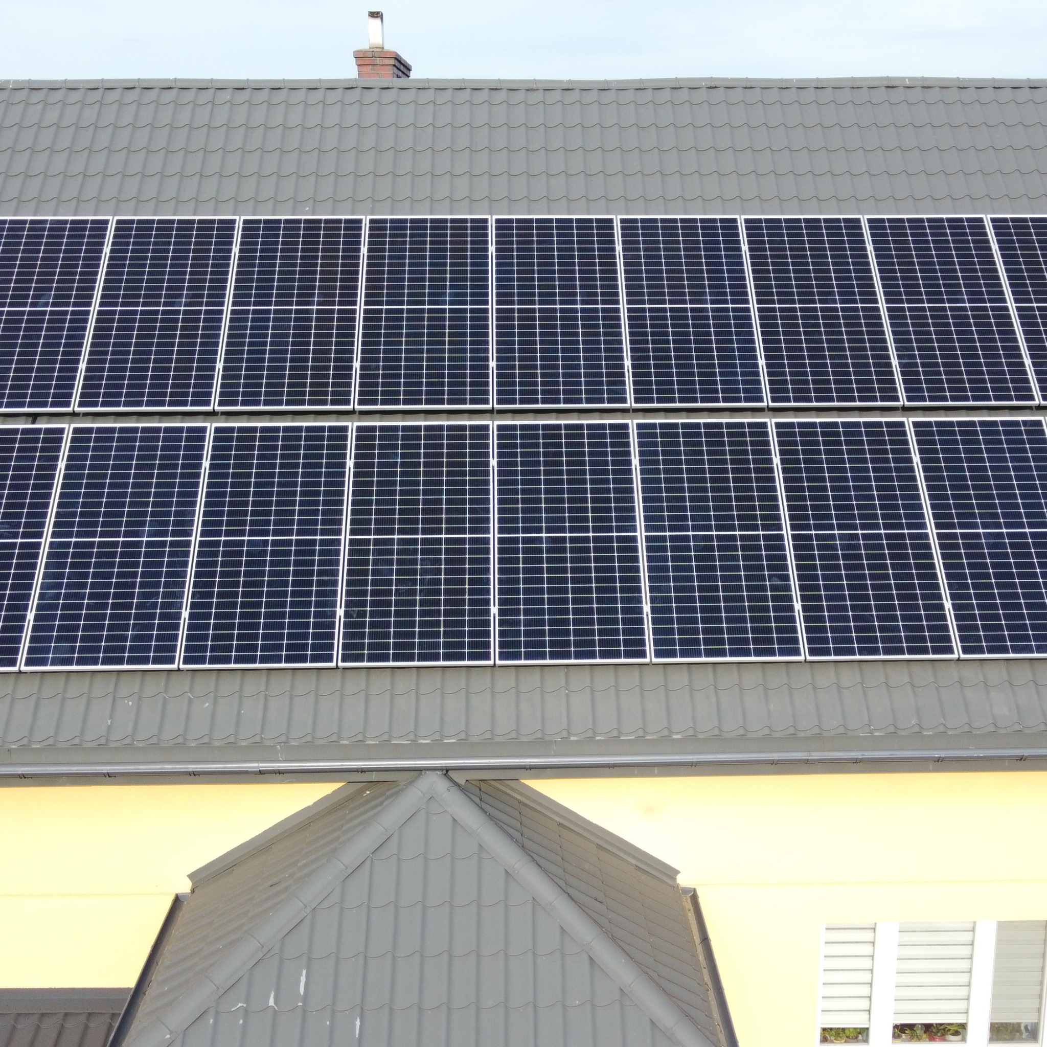 Panele fotowoltaiczne zamontowane na szarym, metalowym dachu domu z ceglanym kominem w tle.