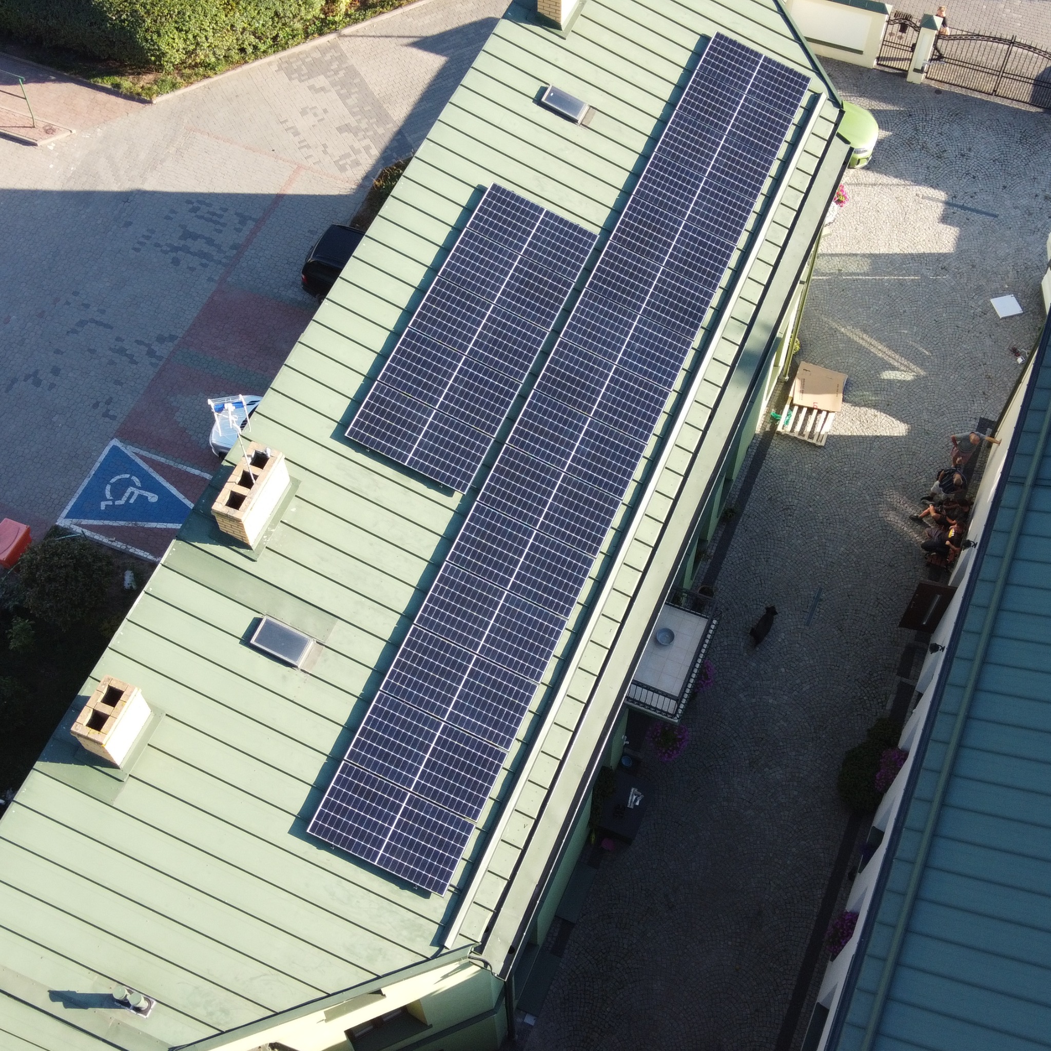 Widok z góry na zielony dach budynku z zainstalowanymi panelami słonecznymi, widoczny fragment parkingu z symbolem osoby niepełnosprawnej oraz wewnętrzny dziedziniec z siedzącymi osobami.