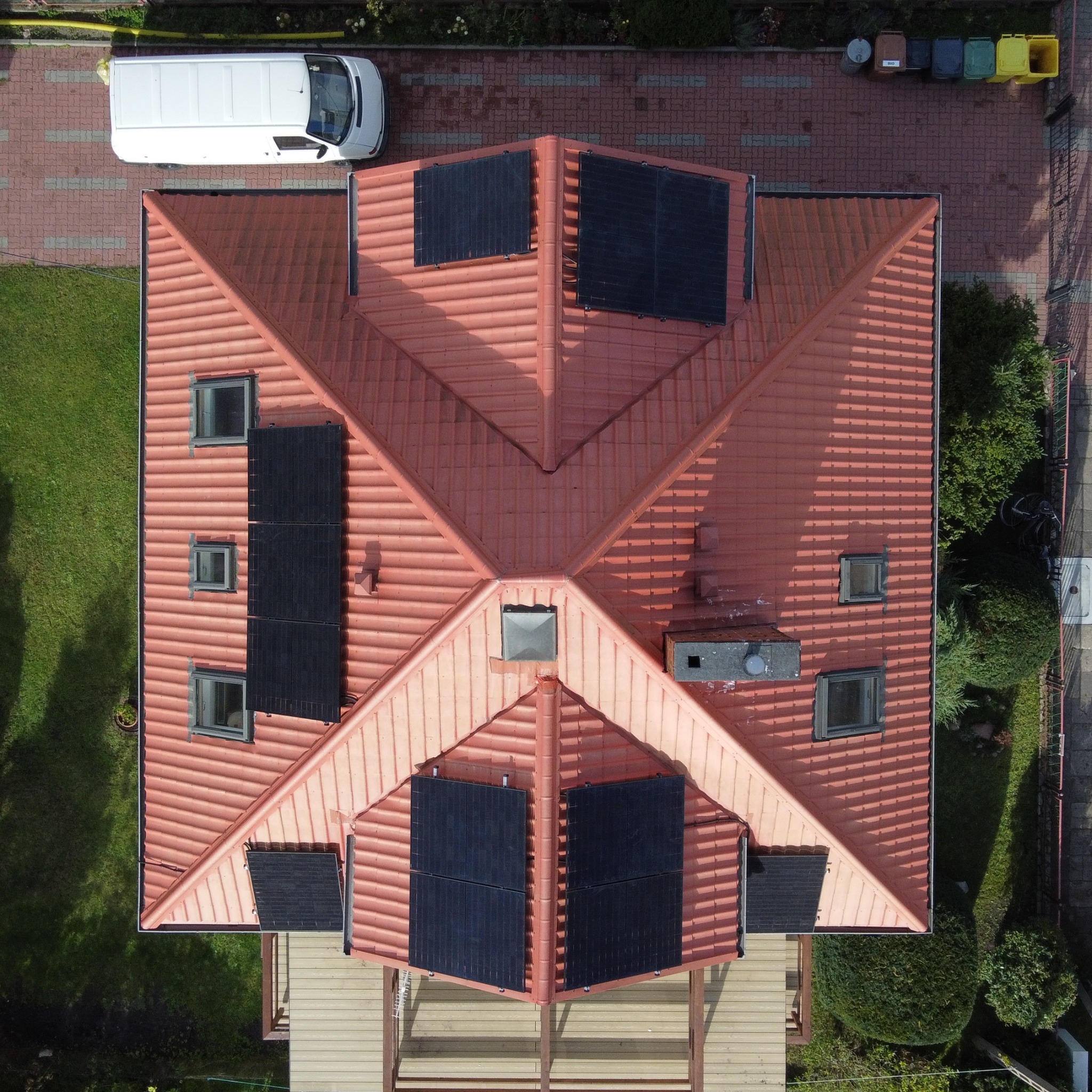 Widok z drona na dach domu z czerwonej dachówki z zamontowanymi panelami słonecznymi, obok zaparkowany biały van i kolorowe pojemniki na śmieci.