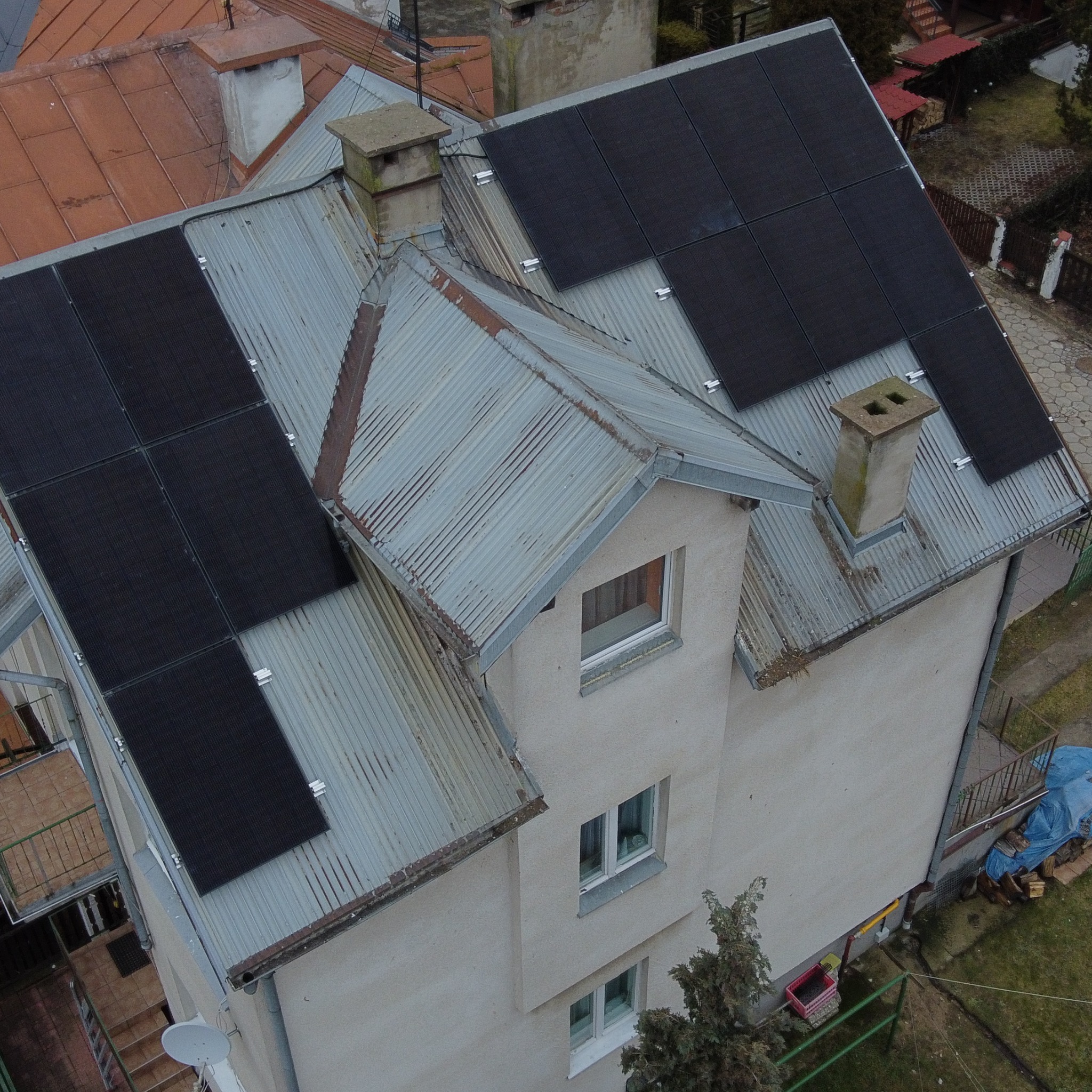 Widok z góry na dach domu z zainstalowanymi panelami fotowoltaicznymi, blaszany dach pokryty patyną, komin z dwoma otworami, otoczenie budynku z zielenią i zabudową.