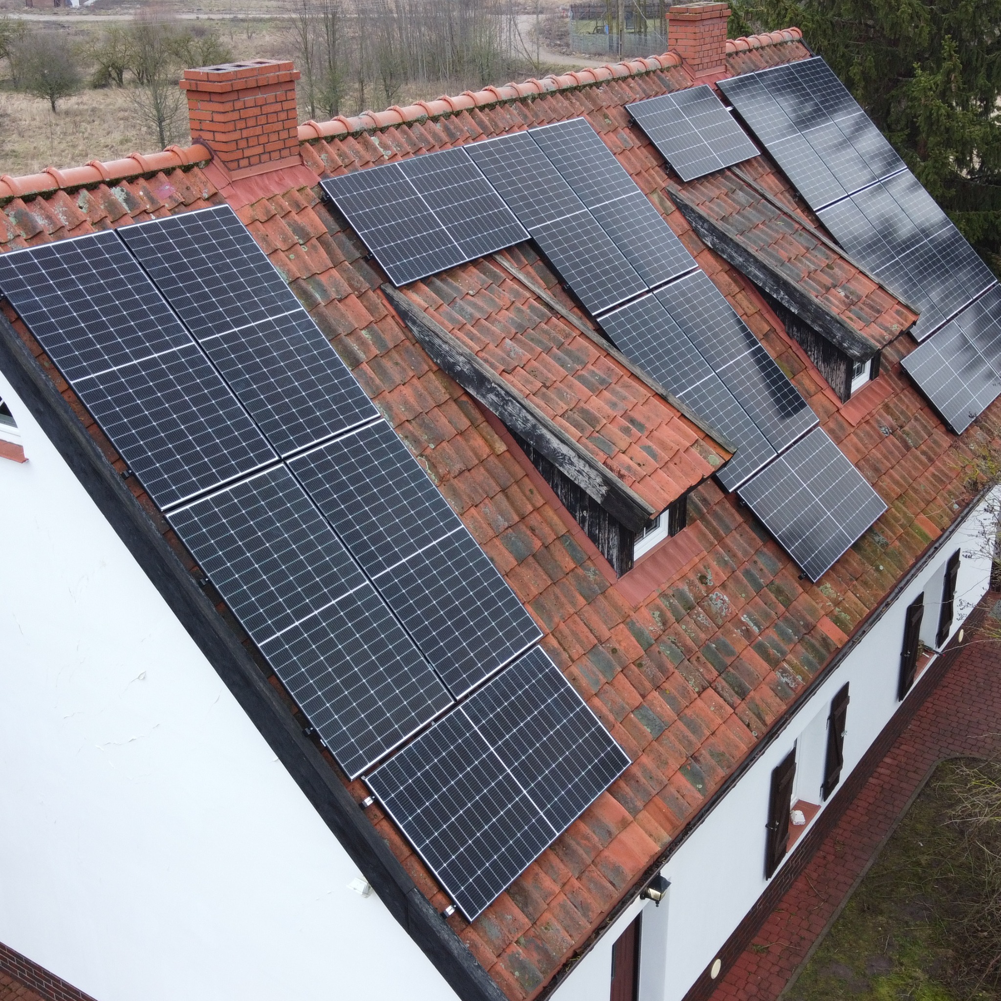 Montaż paneli słonecznych na dachu z czerwonej dachówki, widoczne kominy i okna dachowe, ujęcie z góry.