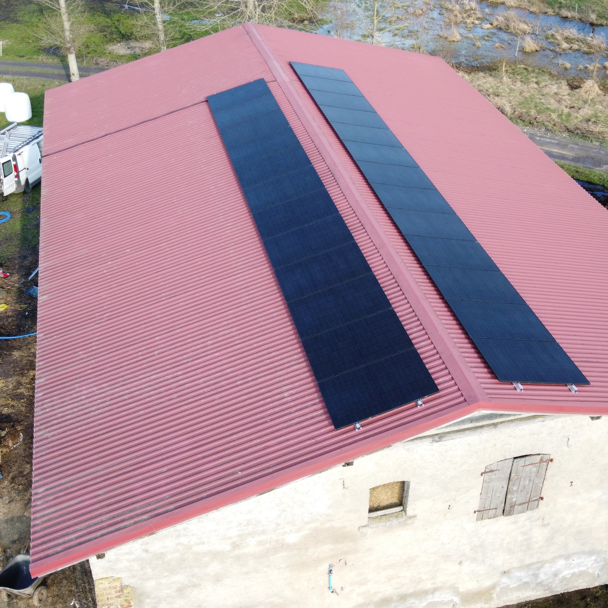 Dwa rzędy paneli słonecznych na czerwonym, blaszany dachu budynku gospodarczego z zasłoniętymi okiennicami, widok z góry, w tle łąka i biały samochód dostawczy.