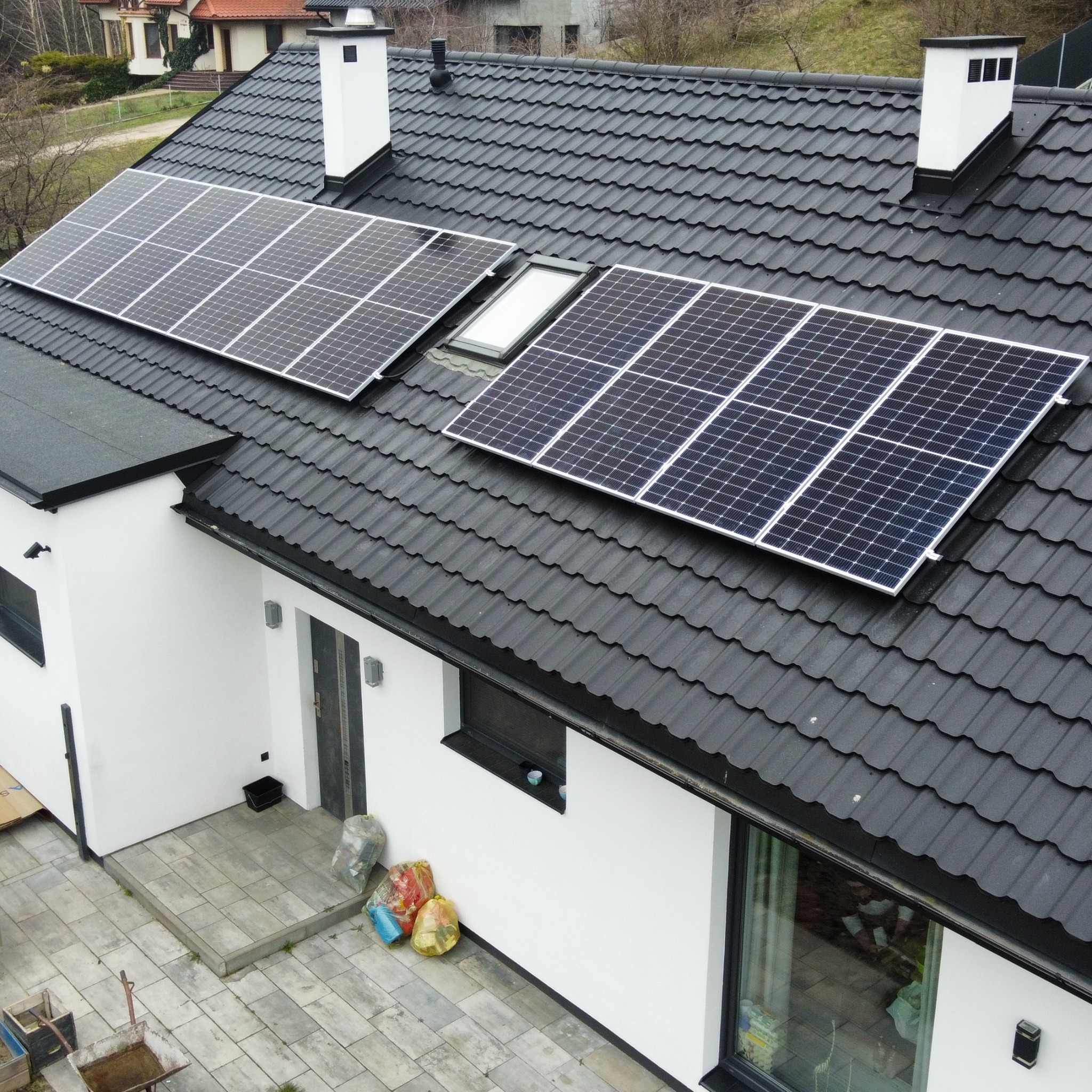 Panele fotowoltaiczne zamontowane na dachu domu z czarną dachówką, widok z góry, w tle inne budynki i drzewa, na dole widoczne worki ze śmieciami.