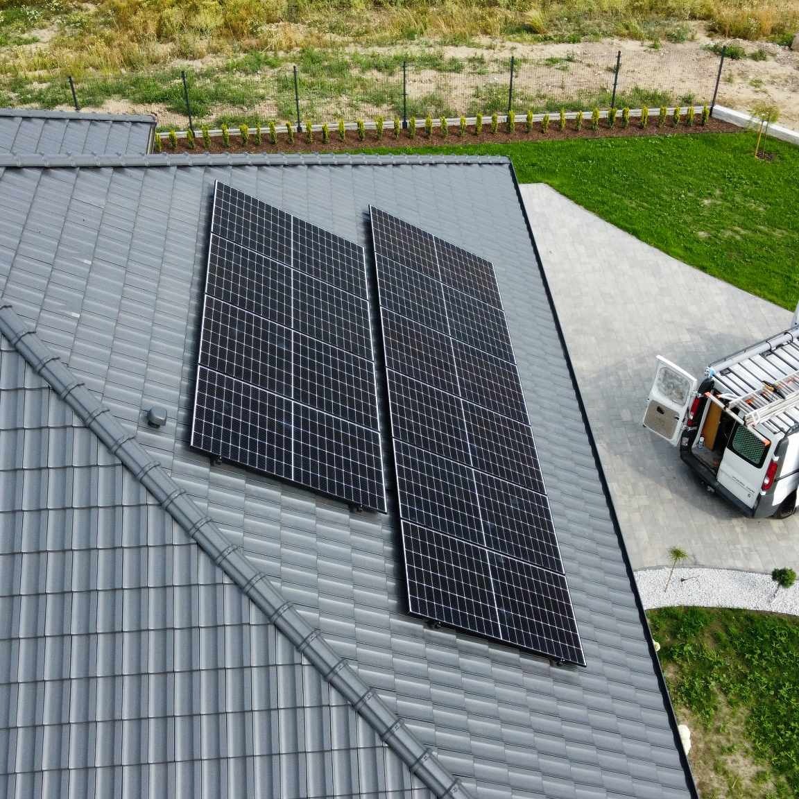 Dwa panele słoneczne zamontowane na szarym, spadzistym dachu z dachówki ceramicznej. W tle widoczny trawnik, ogrodzenie i zaparkowany biały van z otwartymi drzwiami.