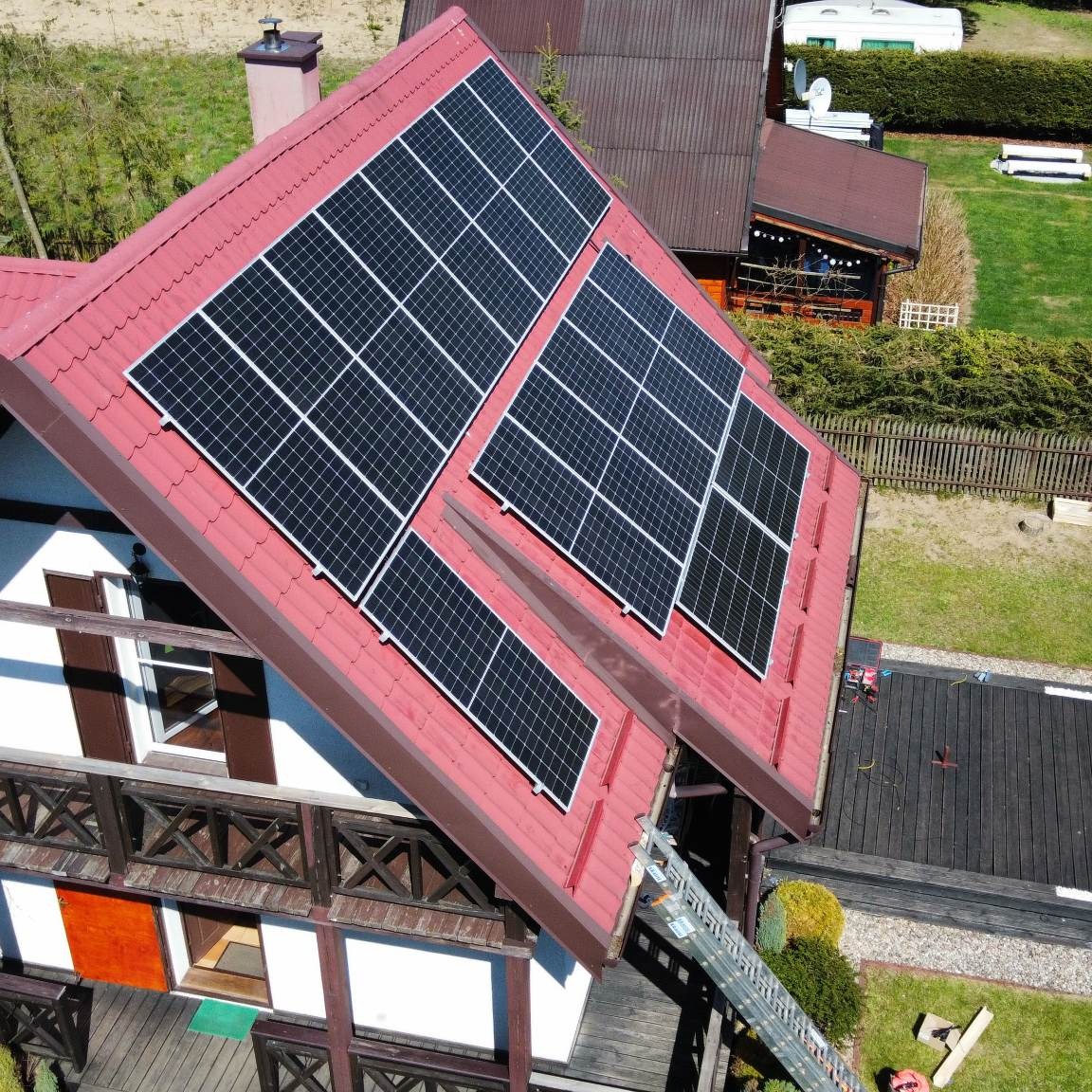 Czerwony dach domu z panelami słonecznymi, widok z góry, drabina oparta o rynnę, drewniany taras obok.