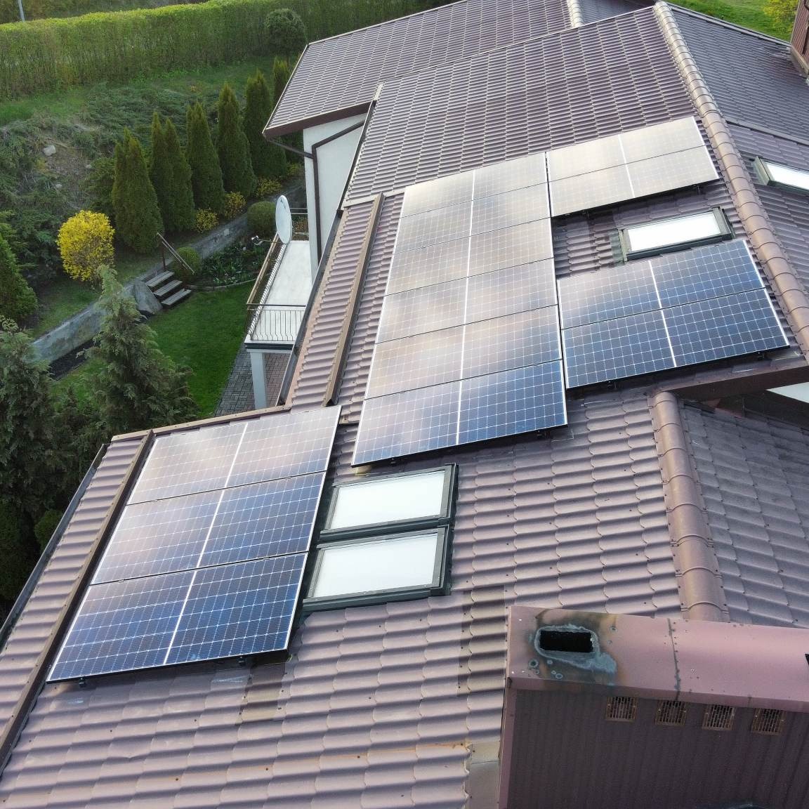 Panele słoneczne na brązowym dachu domu z oknami dachowymi, widok z góry, komin z widocznymi śladami użytkowania.