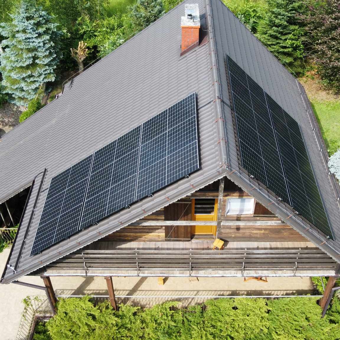 Widok z góry na dom z brązową dachówką i panelami słonecznymi, otoczony zielenią. Widoczny komin i drewniana elewacja pod dachem.