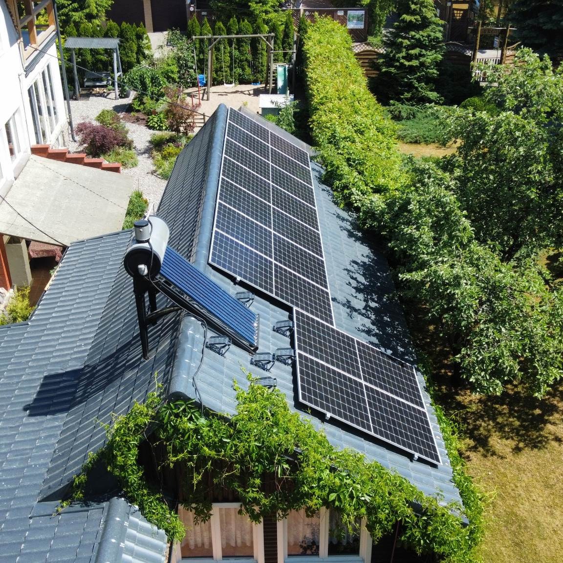Widok z góry na dach domu z panelami fotowoltaicznymi, kolektorem słonecznym i pnącą roślinnością przy oknie.