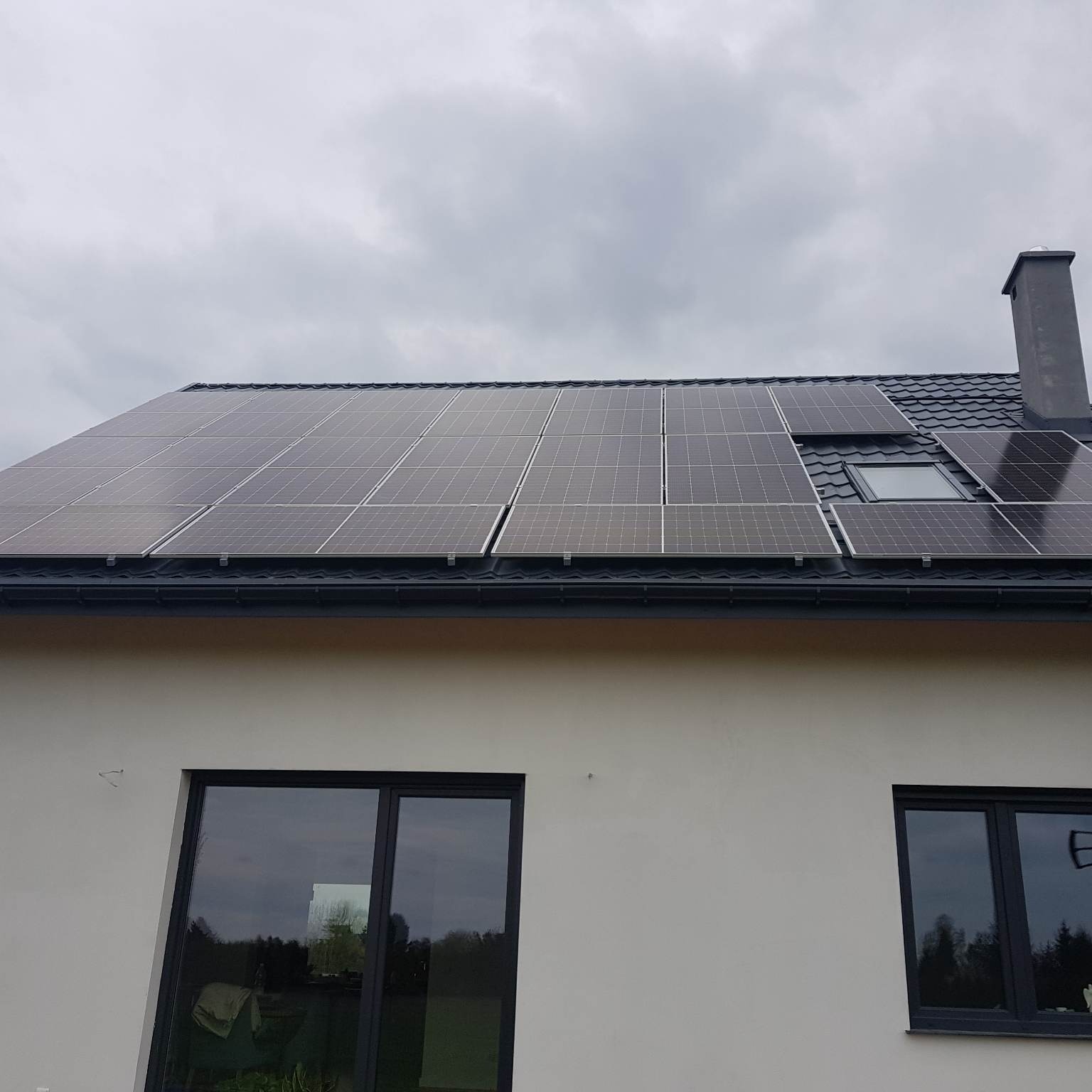 Panele słoneczne na dachu domu jednorodzinnego z czarną dachówką, widoczne również okna dachowe i komin, pochmurne niebo w tle.