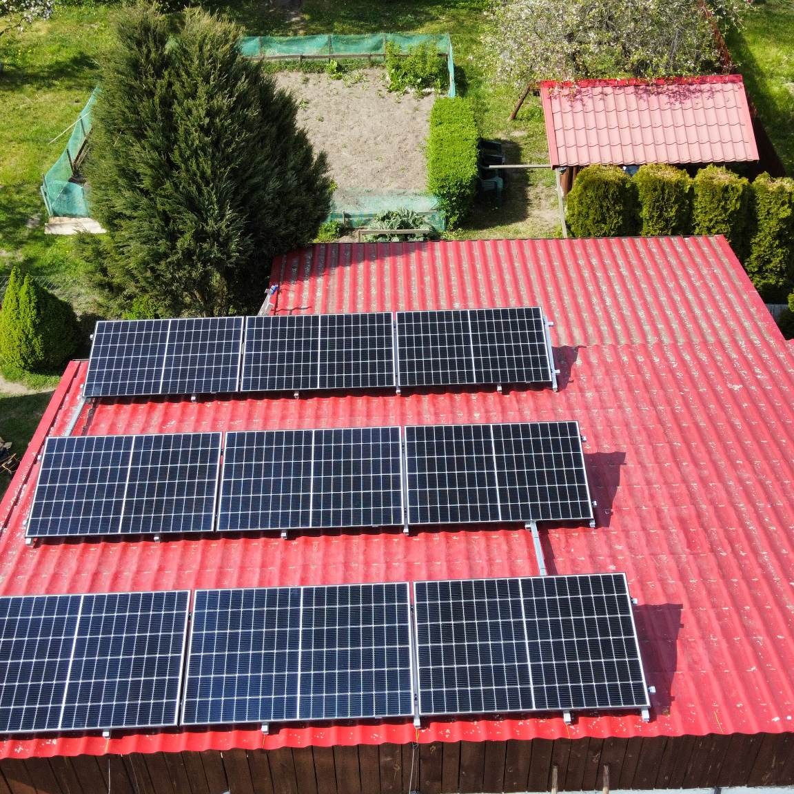 Widok z góry na panele słoneczne zamontowane na czerwonym, falistym dachu budynku, z ogrodem i małą szopą w tle.