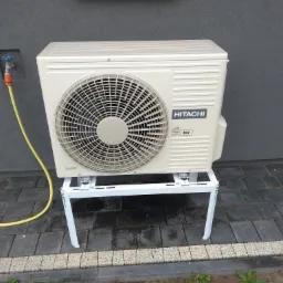 Zewnętrzna jednostka klimatyzacji Hitachi R32 na białym stelażu, przymocowana do szarej ściany budynku z kranem i żółtym wężem ogrodowym.