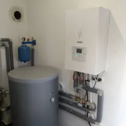 Biała jednostka grzewcza Yutaki zamontowana na ścianie, połączona z szarym zbiornikiem akumulacyjnym i rurami w kotłowni.