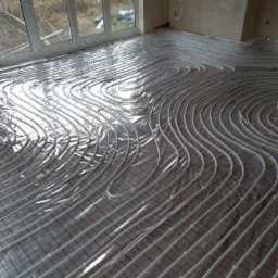 Rury ogrzewania podłogowego ułożone na folii izolacyjnej z siatką, widok na duże okno balkonowe i surowe ściany w tle.