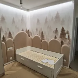 Pokój dziecięcy w trakcie urządzania: białe łóżko z szufladami i drewnianym stelażem, tapeta z motywem leśnym i panelami ściennymi w kształcie drzew, podłoga ułożona w jodełkę, kartony na podłodze.