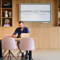 Mężczyzna w okularach siedzi przy stole z laptopem i notatnikiem w nowoczesnym biurze. Na ścianie za nim wisi ekran z logotypem firmy 'Kamiński KF Finanse - Produkty Hipoteczne' oraz regał...