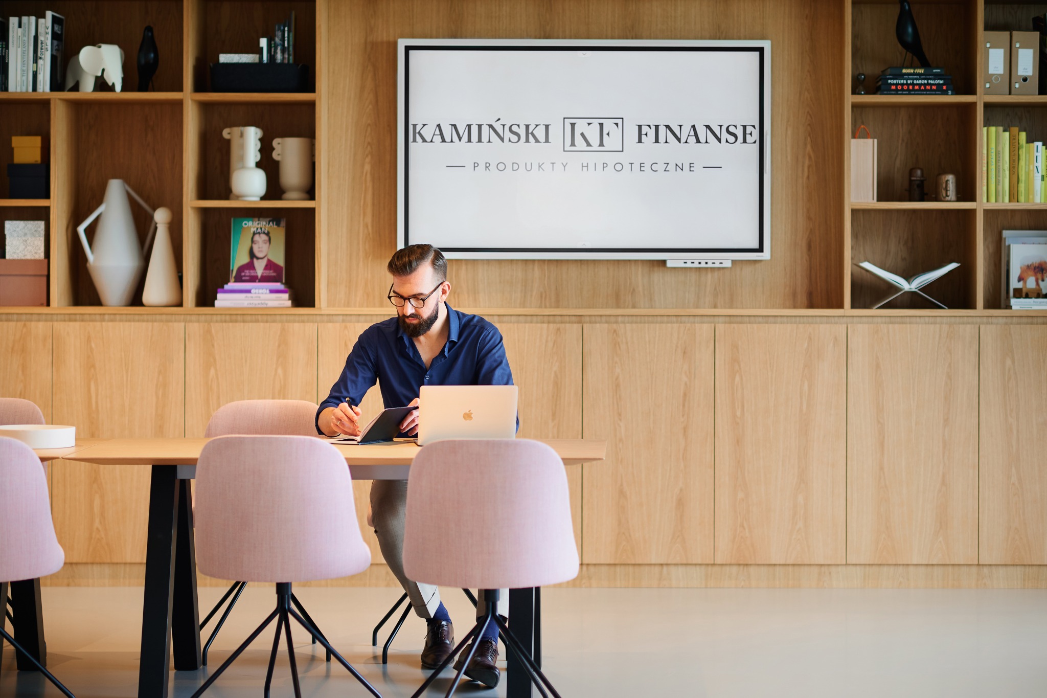 Mężczyzna w okularach siedzi przy stole z laptopem i notatnikiem w nowoczesnym biurze. Na ścianie za nim wisi ekran z logotypem firmy 'Kamiński KF Finanse - Produkty Hipoteczne' oraz regał...