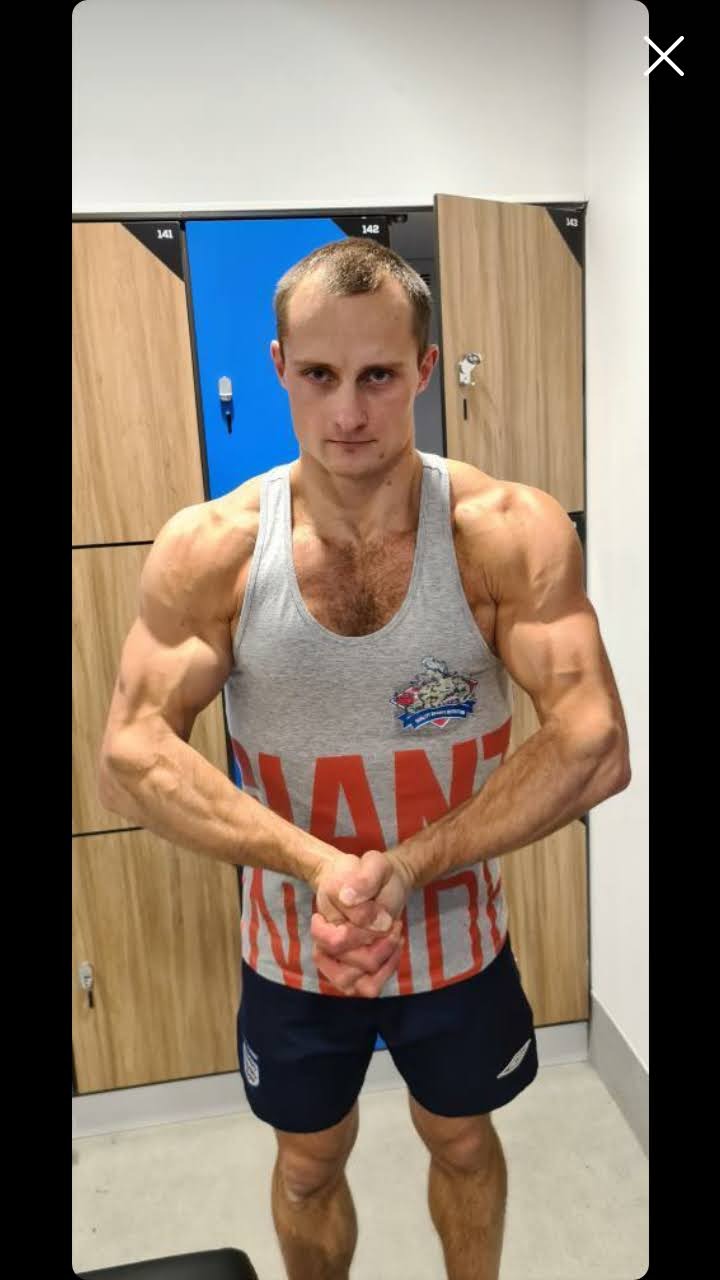 Mężczyzna z napiętymi mięśniami w szarej koszulce z napisem 'GIANTS' prezentuje sylwetkę w szatni z szafkami w tle.