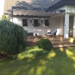 Nowoczesna pergola z rozsuwanym zadaszeniem materiałowym, zadaszająca taras z deskami w kolorze jasnego drewna, z meblami ogrodowymi i roślinami w doniczkach.