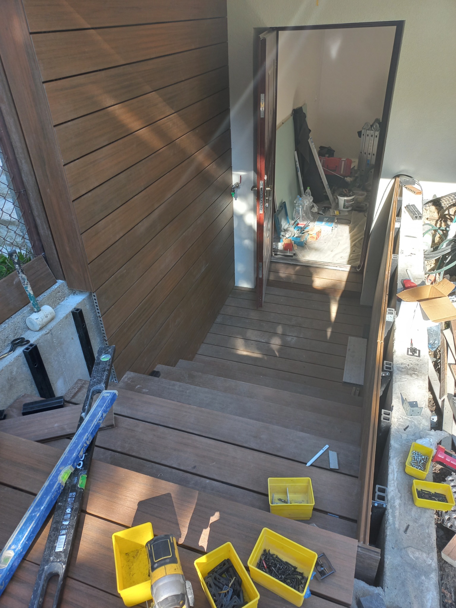 Montaż schodów z desek na zewnątrz budynku, widoczne narzędzia: poziomica, wkrętarka, żółte pojemniki z wkrętami, w tle otwarte drzwi do pomieszczenia z materiałami budowlanymi.