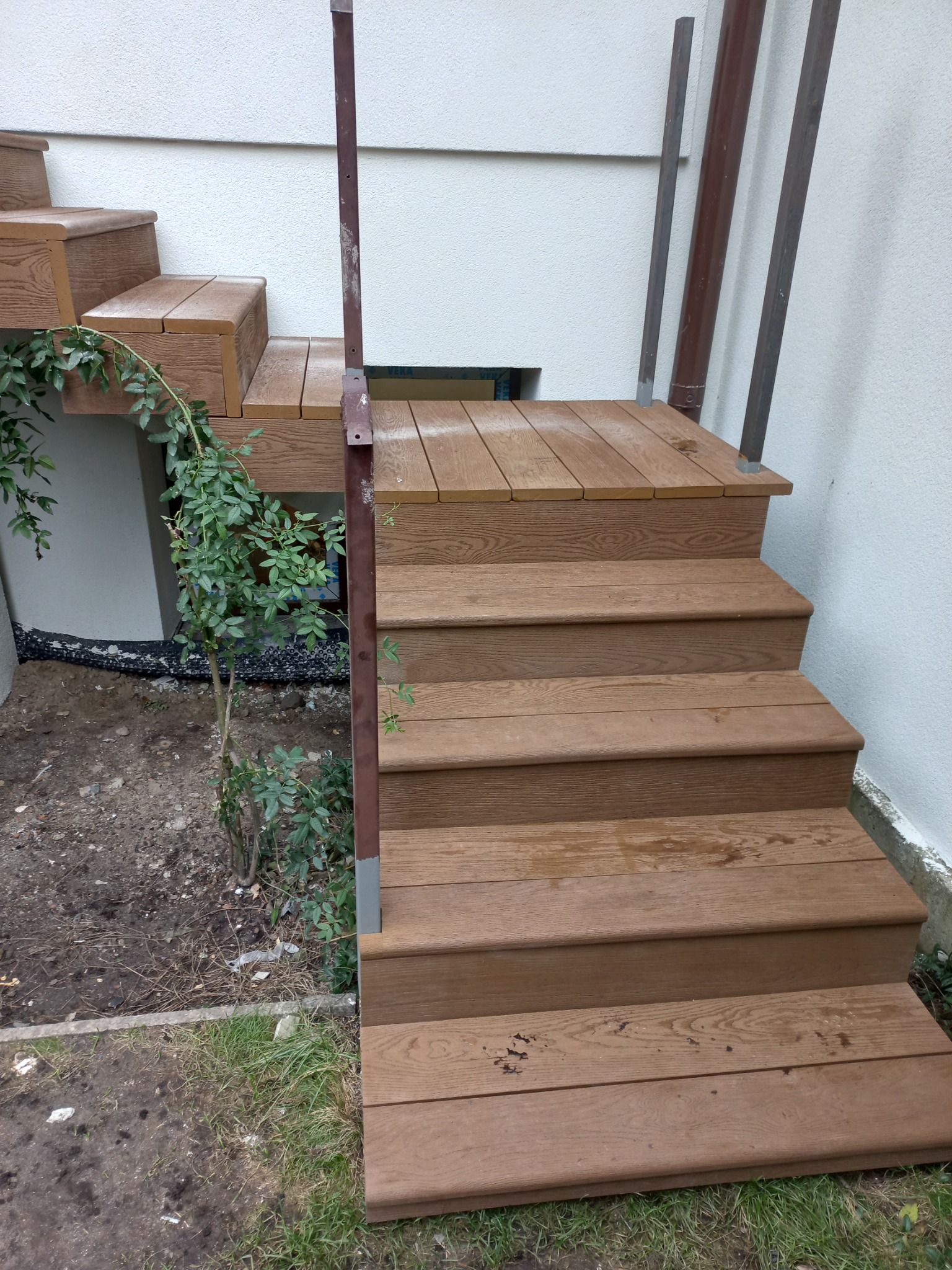 Schody zewnętrzne z brązowych desek, z metalową poręczą, obok rosnące pnącze, widoczne ślady po deszczu na deskach.