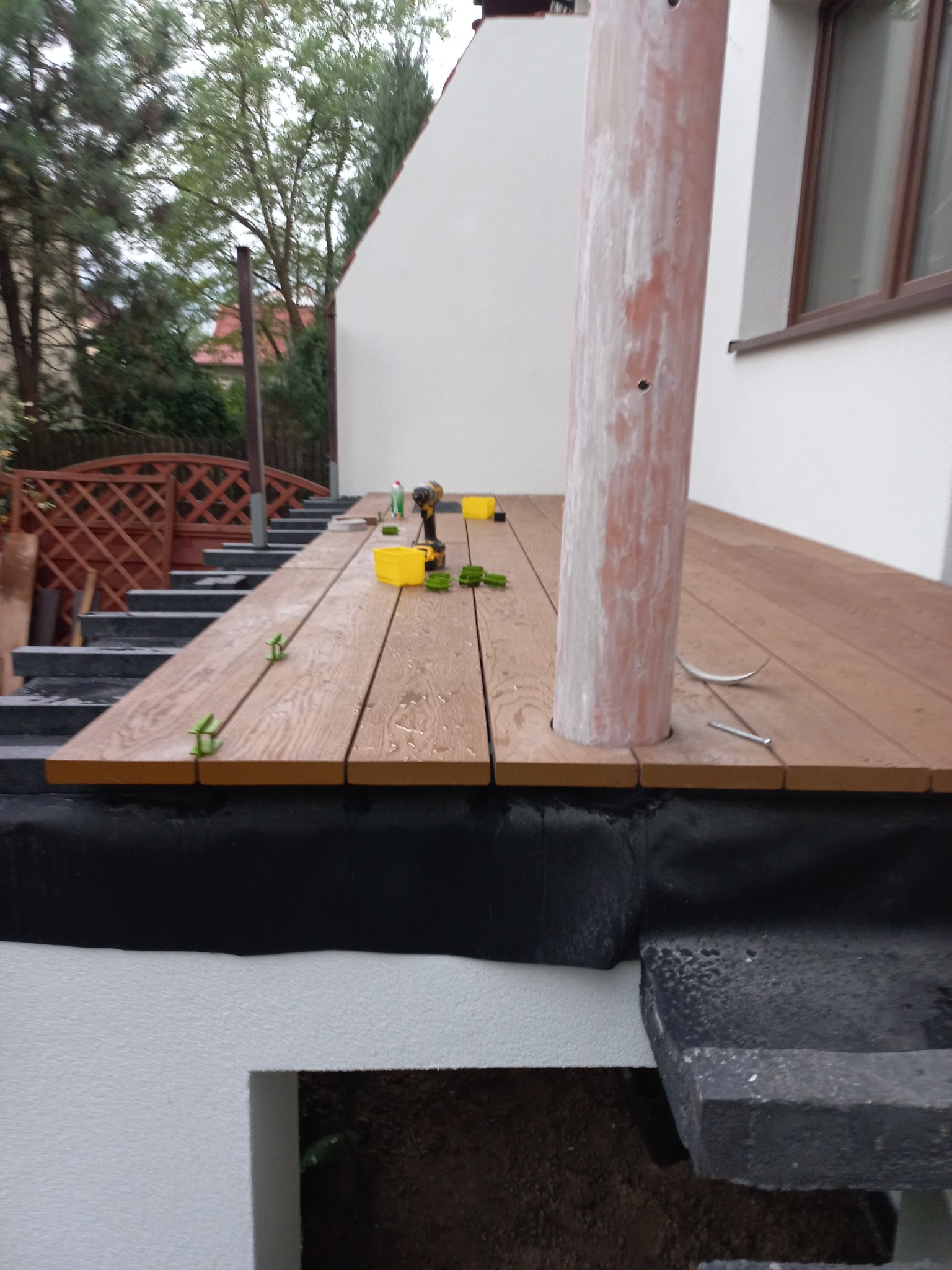 Montaż desek na tarasie, widoczne elementy mocujące, wkrętarka, fragment słupa pergoli i schody prowadzące na taras.