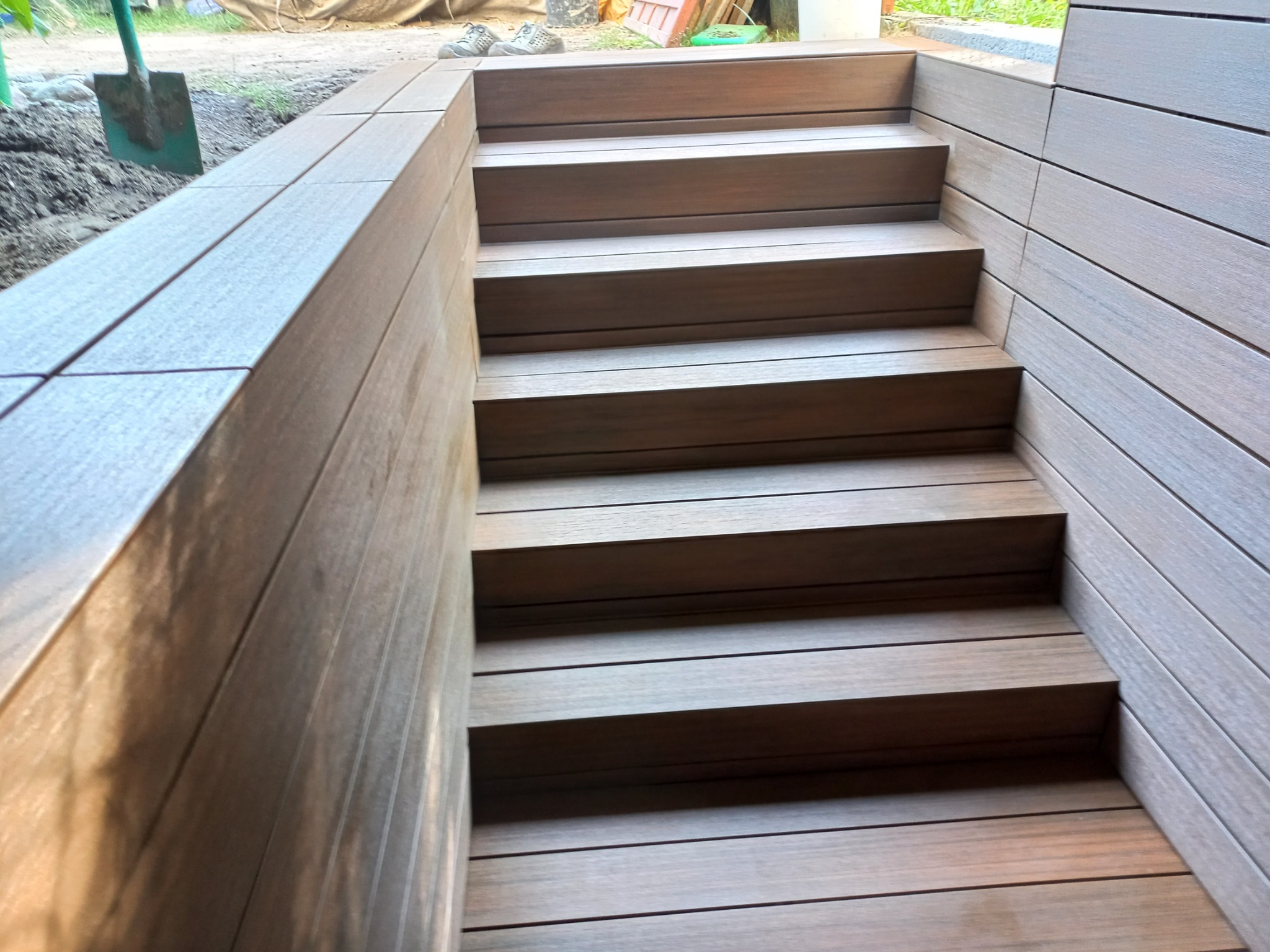 Schody zewnętrzne z desek kompozytowych w kolorze brązowym, prowadzące w dół, widoczne narzędzia ogrodnicze w tle.