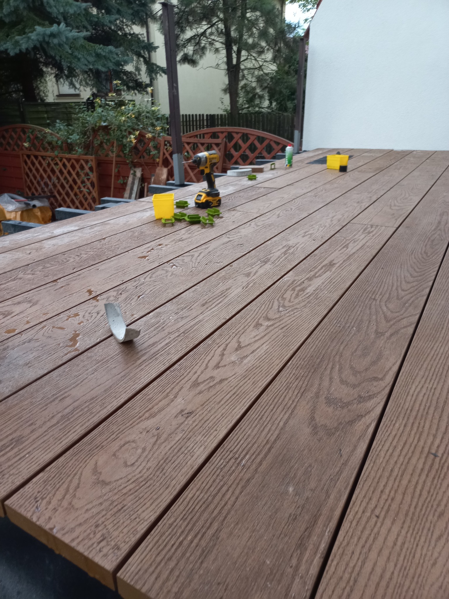 Montaż desek na tarasie: widok na częściowo ułożone deski, wkrętarka DeWalt, pojemnik na wkręty i zielone klipsy montażowe, widoczne elementy konstrukcji pod spodem, tło z zielenią i białym budynkiem.
