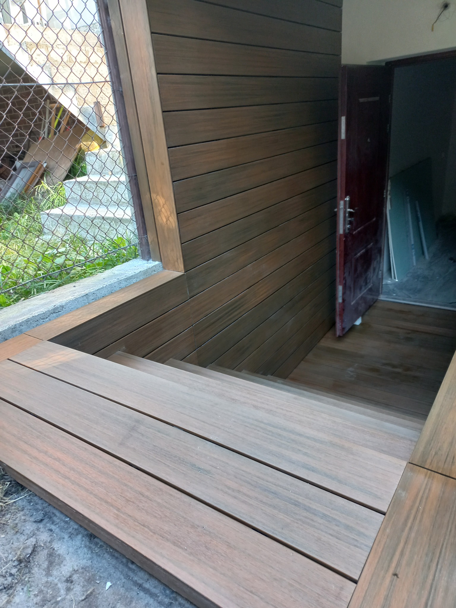 Schody z desek kompozytowych prowadzące do drzwi wejściowych, z widoczną ścianą wykończoną tym samym materiałem i siatką ogrodzeniową w tle.