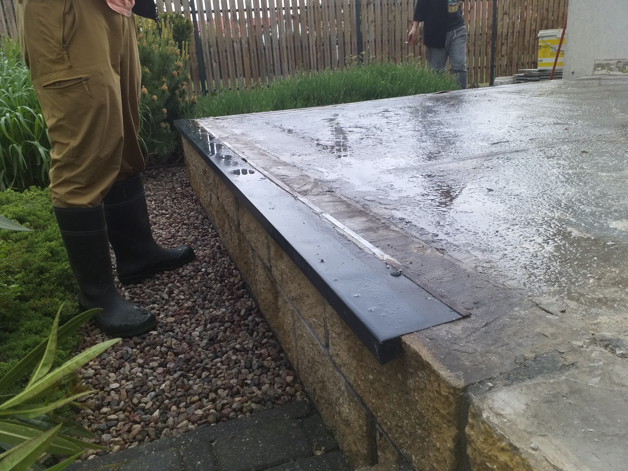 Mokry, betonowy taras w trakcie budowy, z widocznymi elementami wykończeniowymi i pracownikiem w gumowych butach.