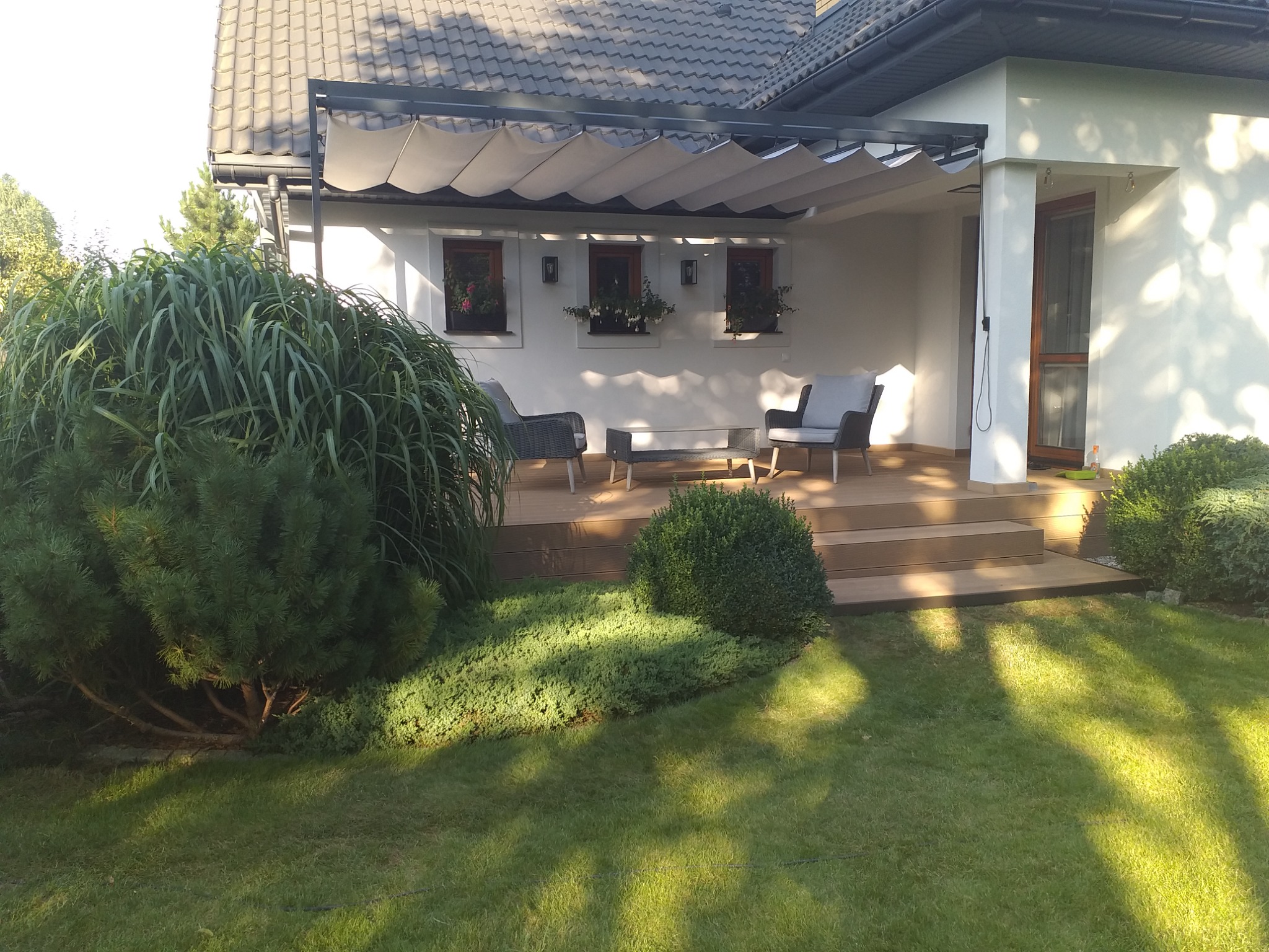 Nowoczesna pergola z rozsuwanym zadaszeniem materiałowym, zadaszająca taras z deskami w kolorze jasnego drewna, z meblami ogrodowymi i roślinami w doniczkach.