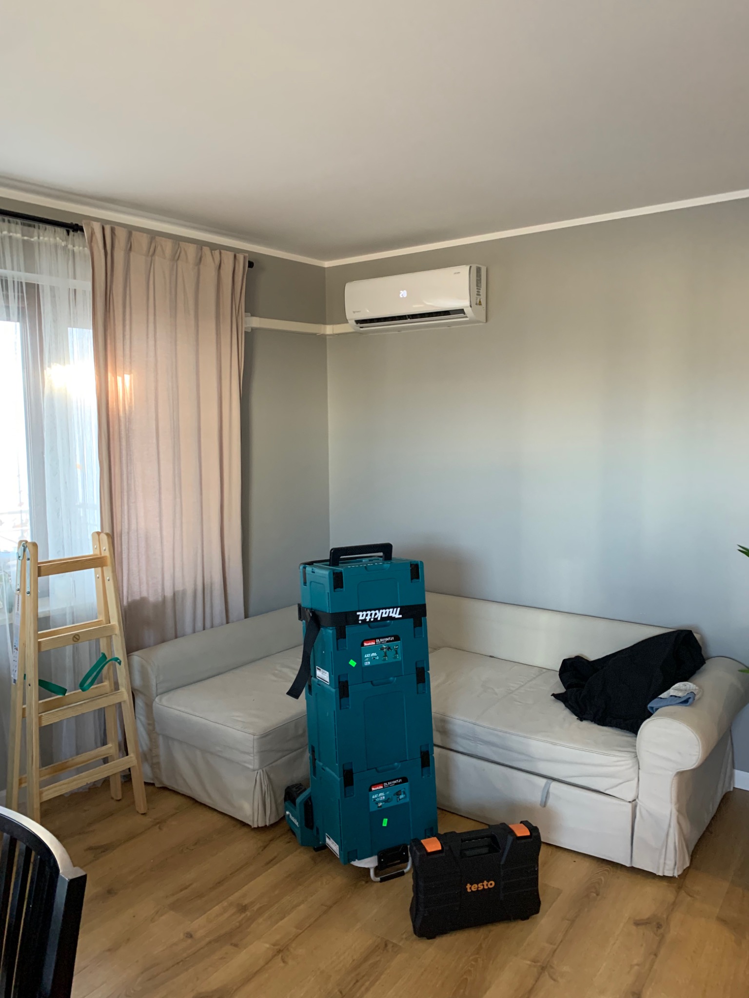 Biały klimatyzator zamontowany na szarej ścianie w pokoju, obok stoi drabina, na podłodze skrzynki narzędziowe Makita i Testo, a pod ścianą biała kanapa.