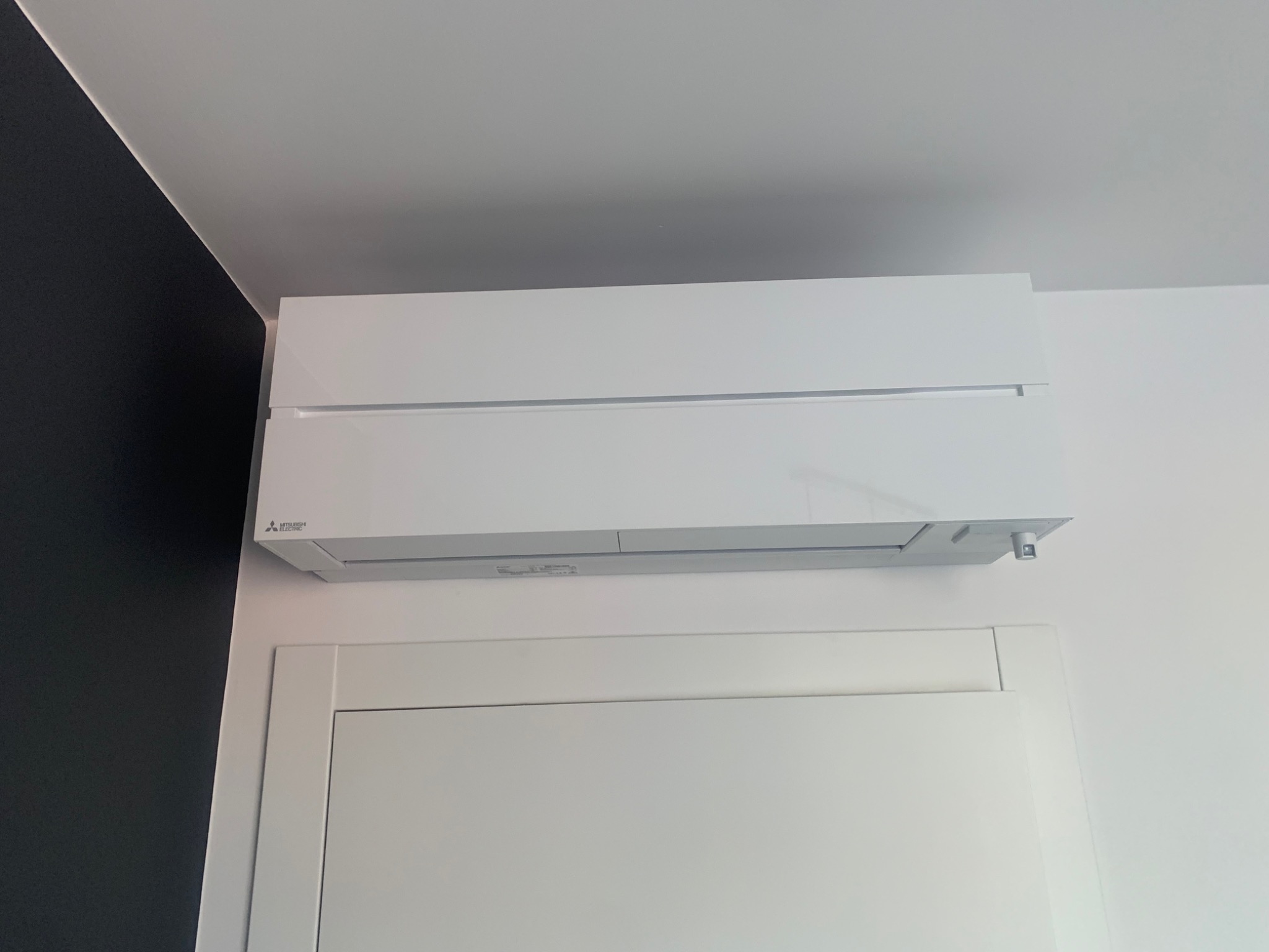 Nowoczesny, biały klimatyzator Mitsubishi Electric zamontowany na ścianie nad białą szafką, z fragmentem czarnej ściany po lewej stronie.