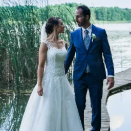 Para młoda w Wolsztynie spaceruje po drewnianym pomoście nad jeziorem, panna młoda w białej sukni z koronkowymi detalami i welonem, pan młody w niebieskim garniturze z butonierką, oboje patrzą...