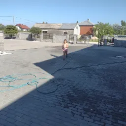 Dziewczynka biegnie po nowo ułożonej, szarej kostce brukowej na podjeździe, widoczny błękitny wąż ogrodowy i ogrodzenie z kamiennymi słupkami i metalowymi przęsłami.