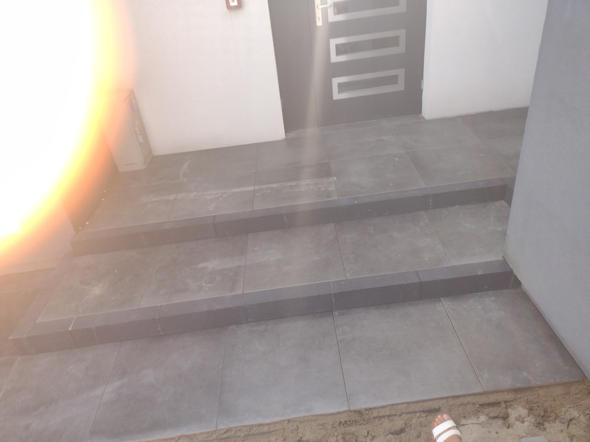 Wykończone, trzy-stopniowe schody wejściowe z szarych płytek, prowadzące do nowoczesnych drzwi wejściowych z metalowymi zdobieniami, z widocznym fragmentem stopy w sandałach w prawym dolnym rogu...